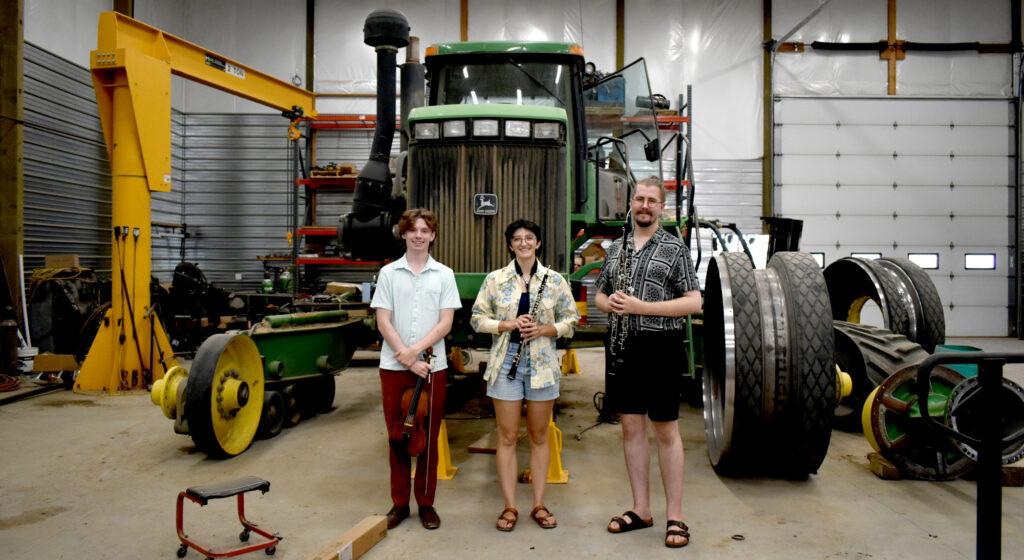 A chamber music trio standing in front of a large tractor in a shop.