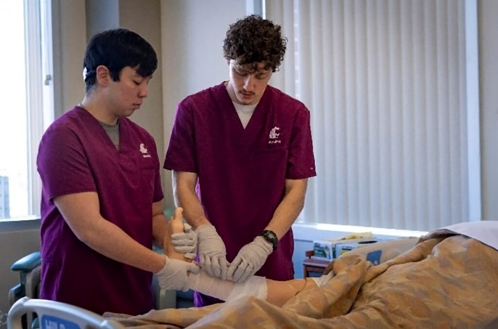 Two male nursing students practice bandaging a leg on a medical training dummy.