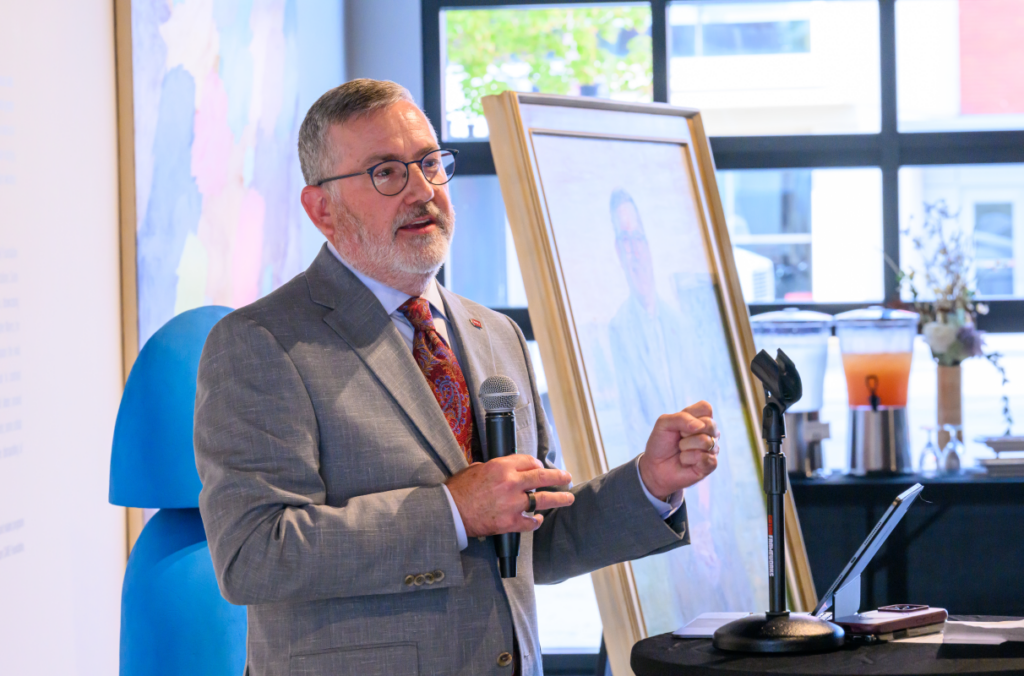 Kirk Schulz speaking in front of portrait.