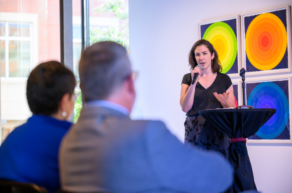 Artist Grace Flott speaks at podium in front of Kirk and Noel Schulz.