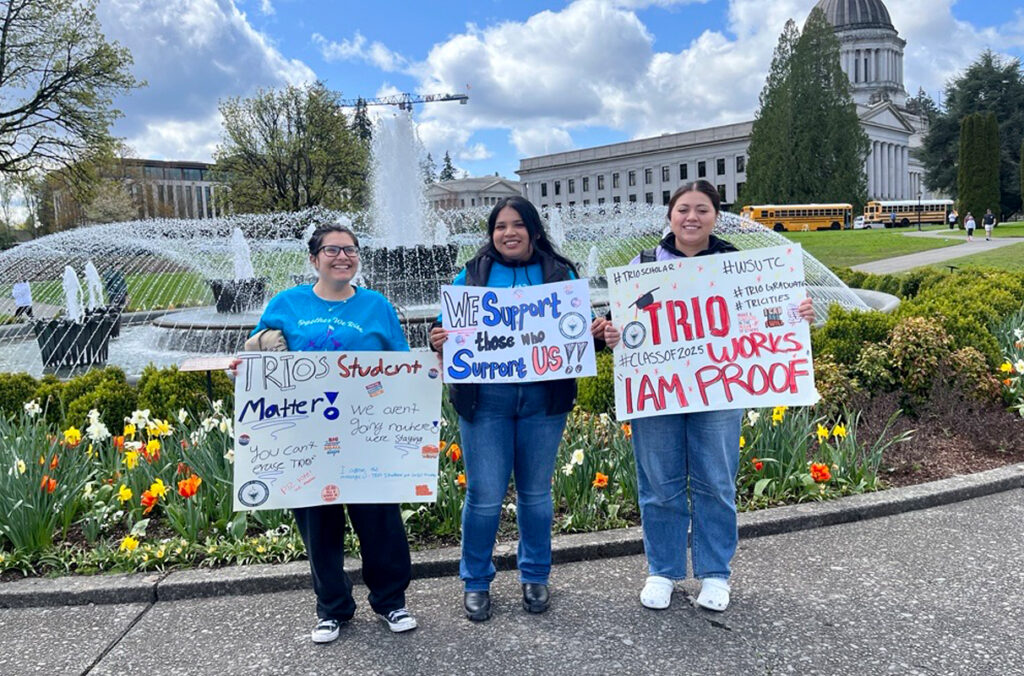 Three students holding signs in support of TRIO in front of a fountain at the Washington State Capitol.