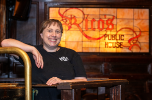 Tawny Szumlas in front of a stained-glass window that reads "Rico's Public House".