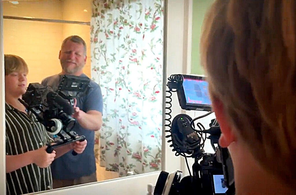 A mentor assisting a teenager holding a video camera in front of a mirror.