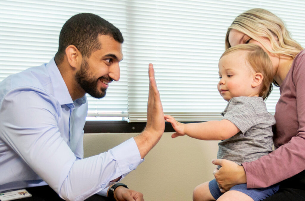 A doctor holds his hand up to high five a toddler sitting on his mother's lap.
