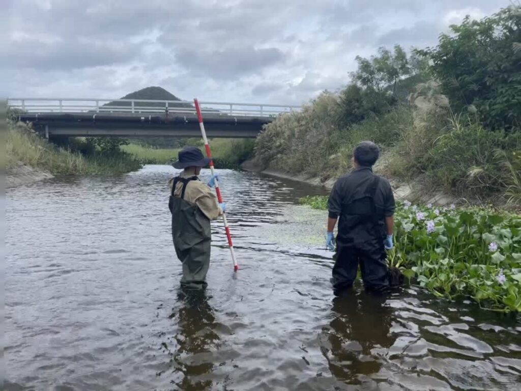 Two Japanese workers stand in water holding tools next to a bridge overpass.