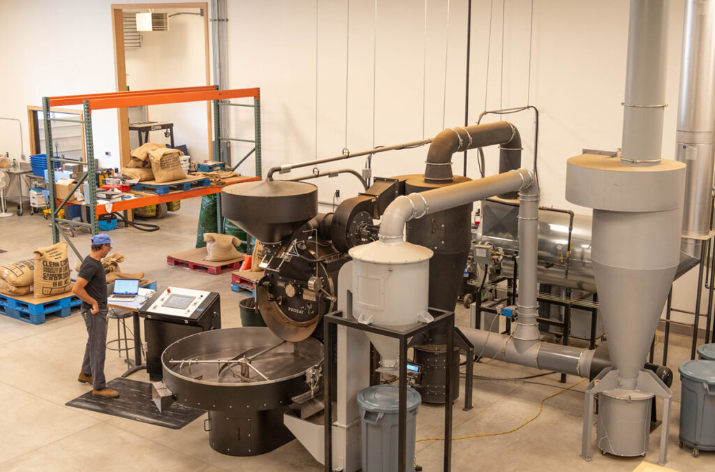 A man standing at the controls of an industrial coffee roasting machine inside a warehouse.