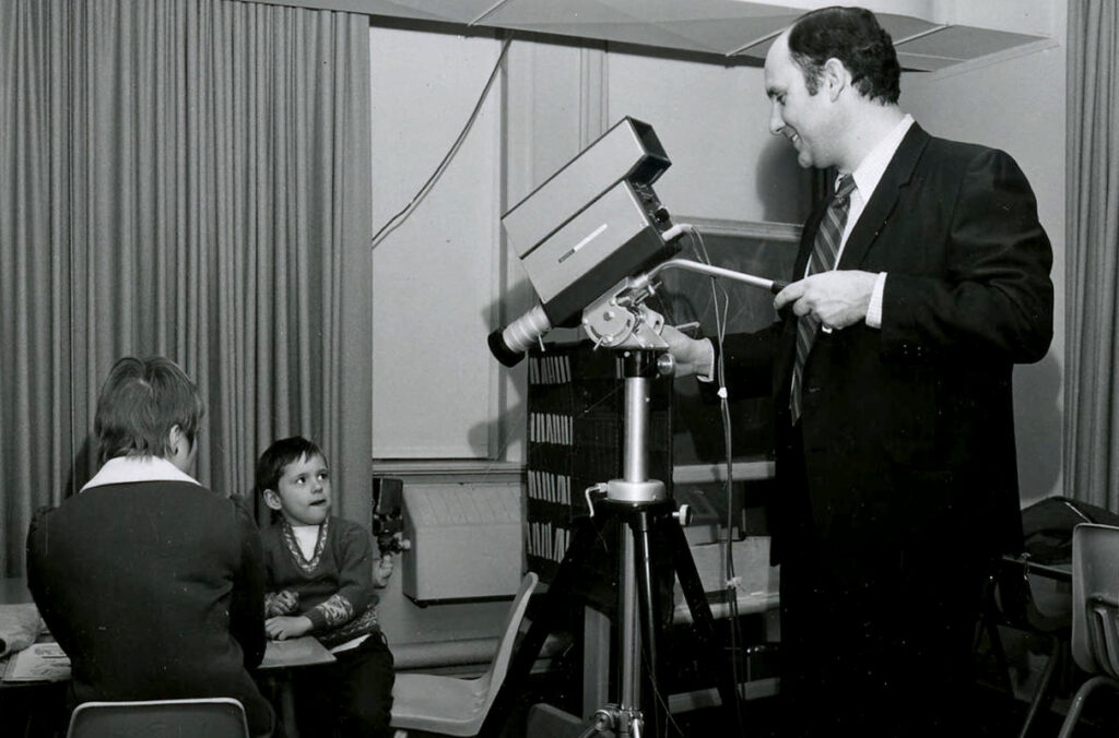 A faculty member videotapes a student clinician working with a child in a black and white photo from 1970.