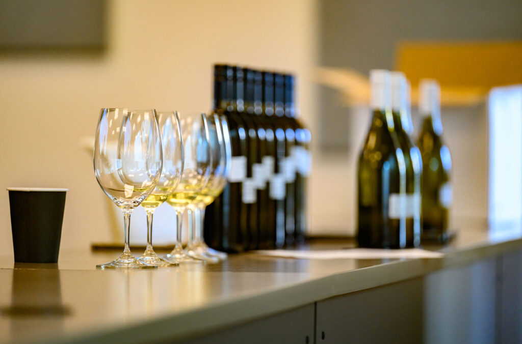 A line of wine glasses for tasting sits next to several bottles of wine on a table.