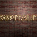 Neon tube sign that reads "Hospitality" against a brick wall.