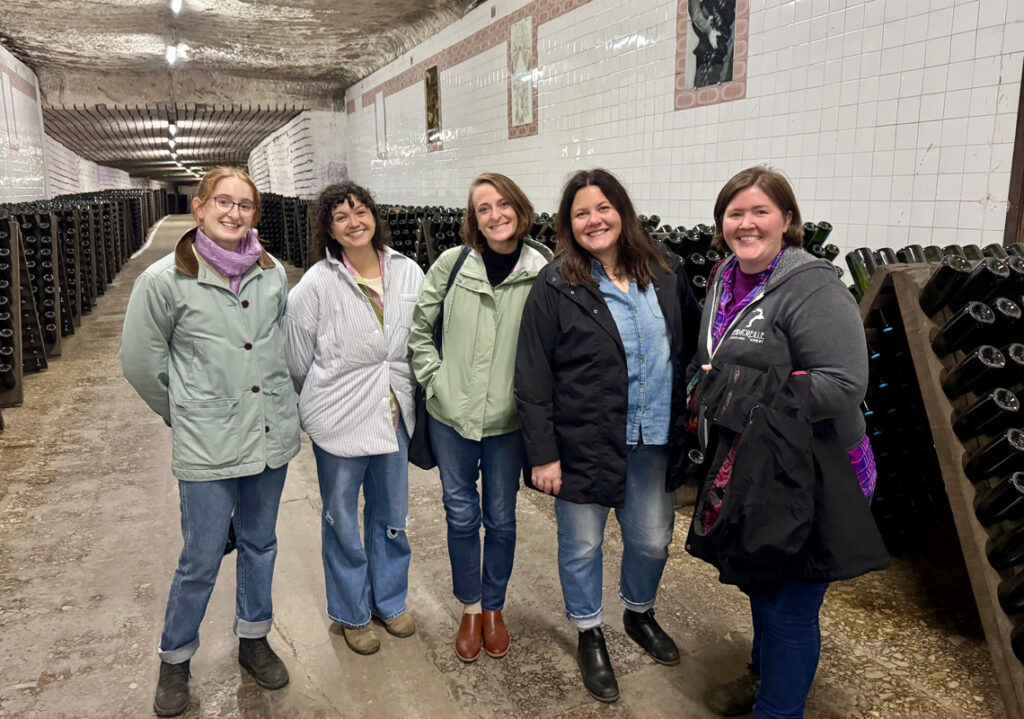 Five women posing for a group photo in a long hallway filled with racks of Moldovan wine.