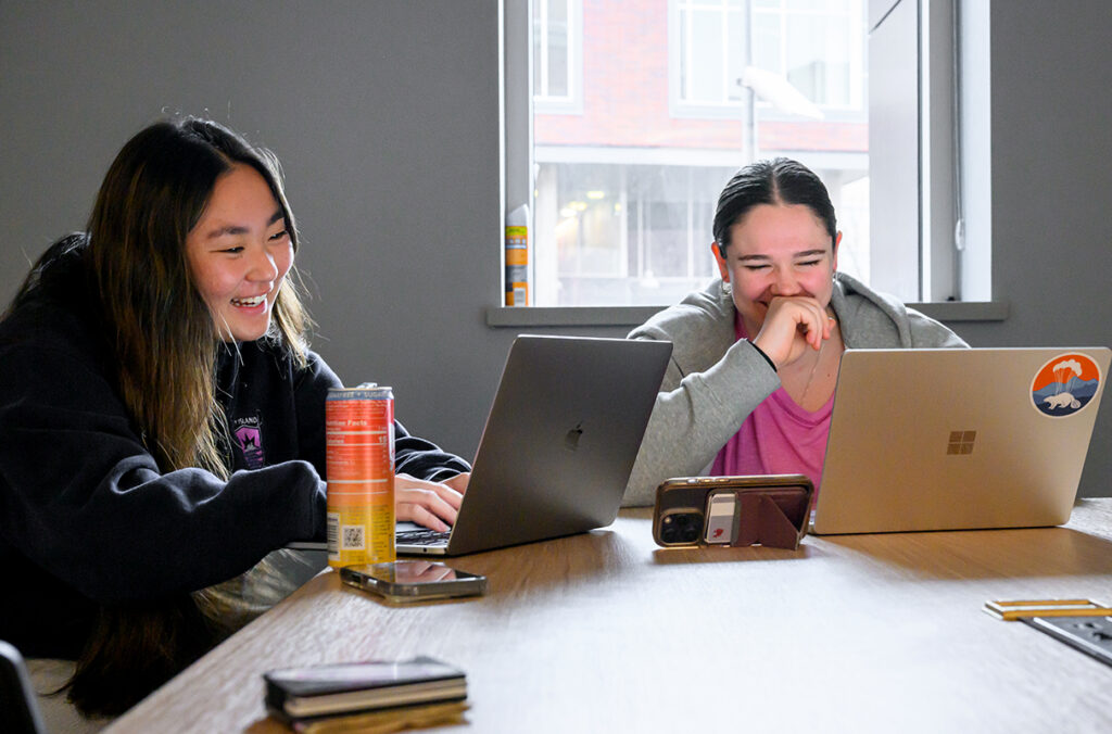 Two WSU student-athletes laughing and working on their laptops at a table.