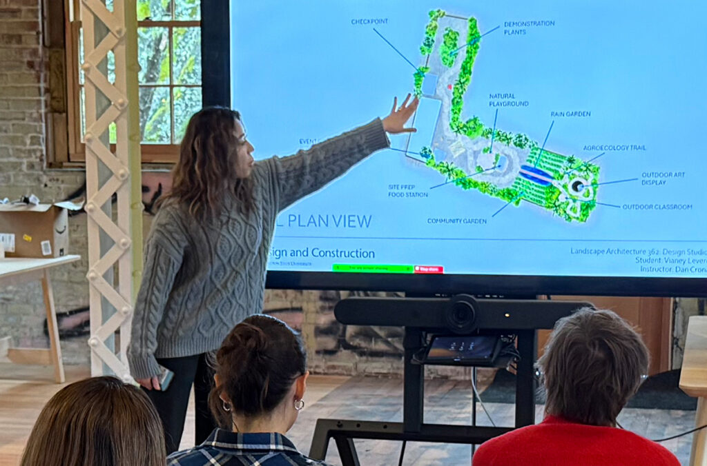 A WSU student gestures towards a digital screen as she presents a design for a Liberian “food hub”.