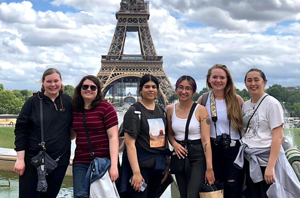Six students pose together in front of the Eiffel Tower, with blue skies and large white clouds overhead.