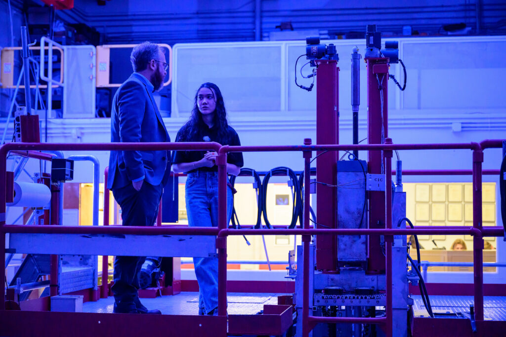 Two people talking in a blue-lit nuclear reactor room surrounded by equipment.