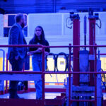 Two people talking in a blue-lit nuclear reactor room surrounded by equipment.