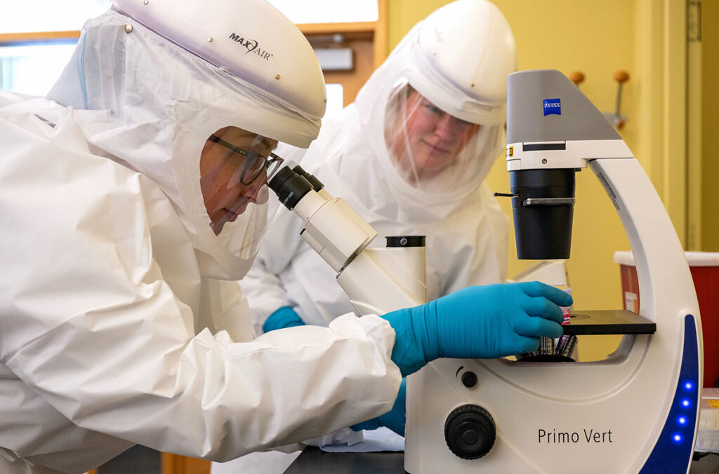 Two researchers wearing full-body PPE as one looks in a microscope.