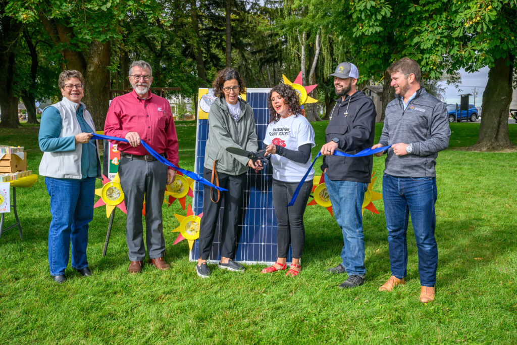 Six people cutting a ribbon in front of a solar panel.