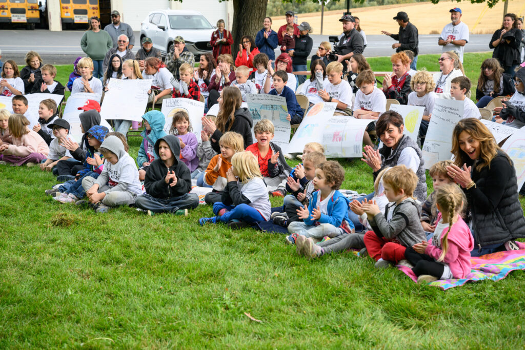 School children sitting in grass clapping with teachers and parents.