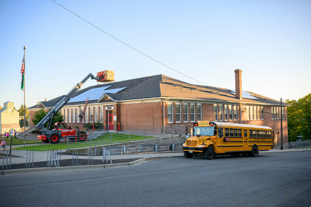 A building with partially installed solar panels on the roof, a crane holding a box, and a school bus.