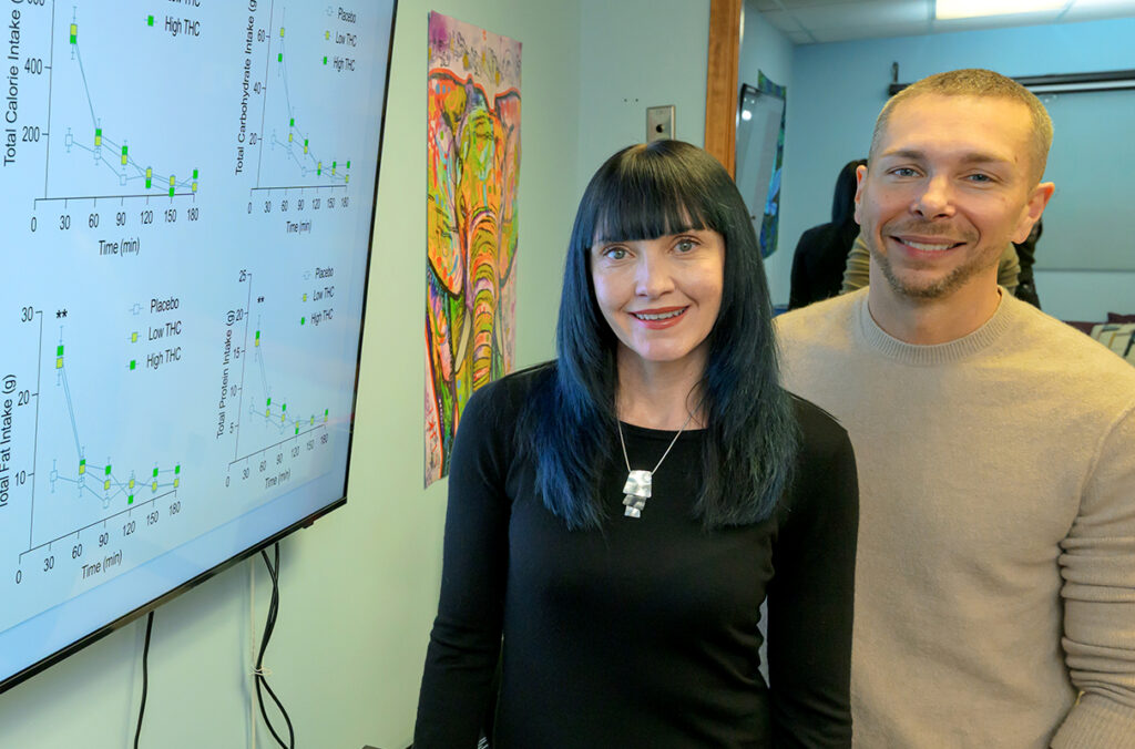 Carrie Cuttler and Ryan McLaughlin stand next to a screen displaying data about caloric intake and THC.