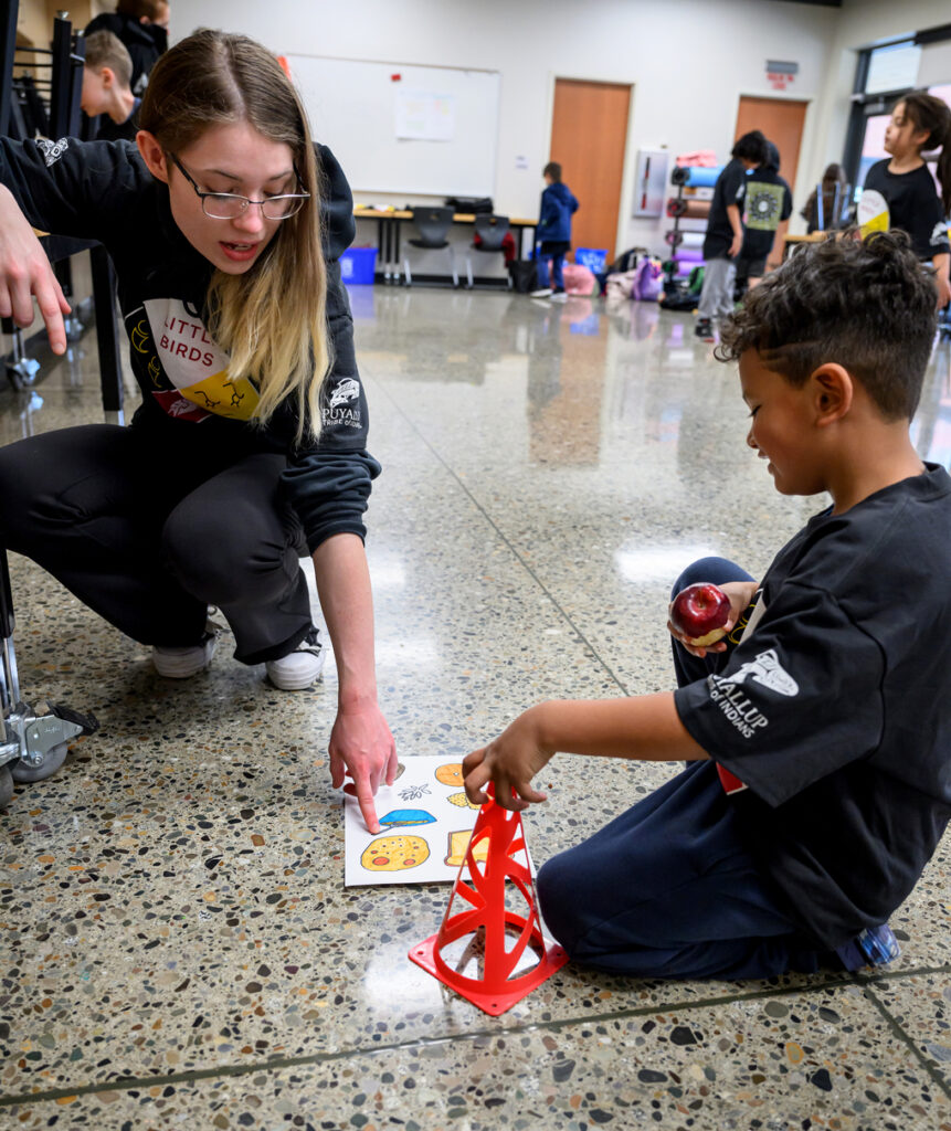 A WSU student points at a sheet describing food groups as a Little Bird student holds a half-eaten apple.