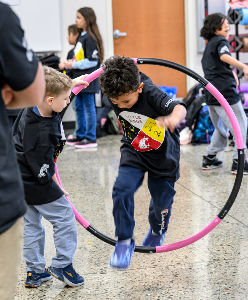 A Little Bird student holding a hula hoop in the air as another student jumps through it.