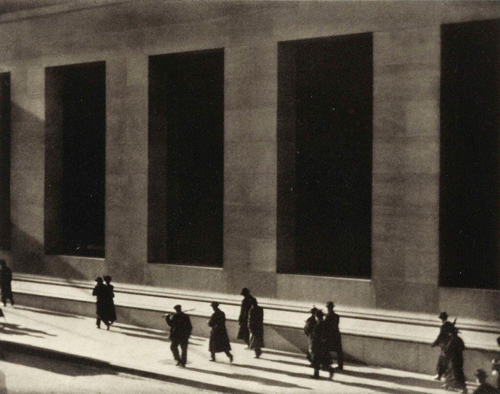 Grainy black-and-white photo of people walking past a large building with tall, dark windows.