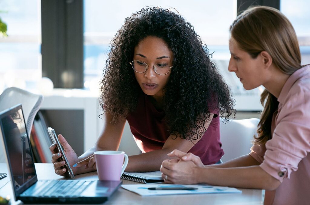 Two women sitting at a desk with a computer. One is pointing at her smartphone as they discuss a news story.