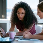 Two women sitting at a desk with a computer. One is pointing at her smartphone as they discuss a news story.