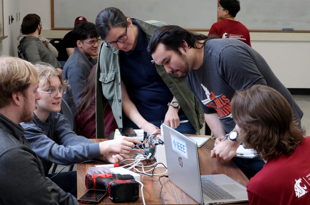 Five hackathon participants sit and stand around a table while collaborating on a project.