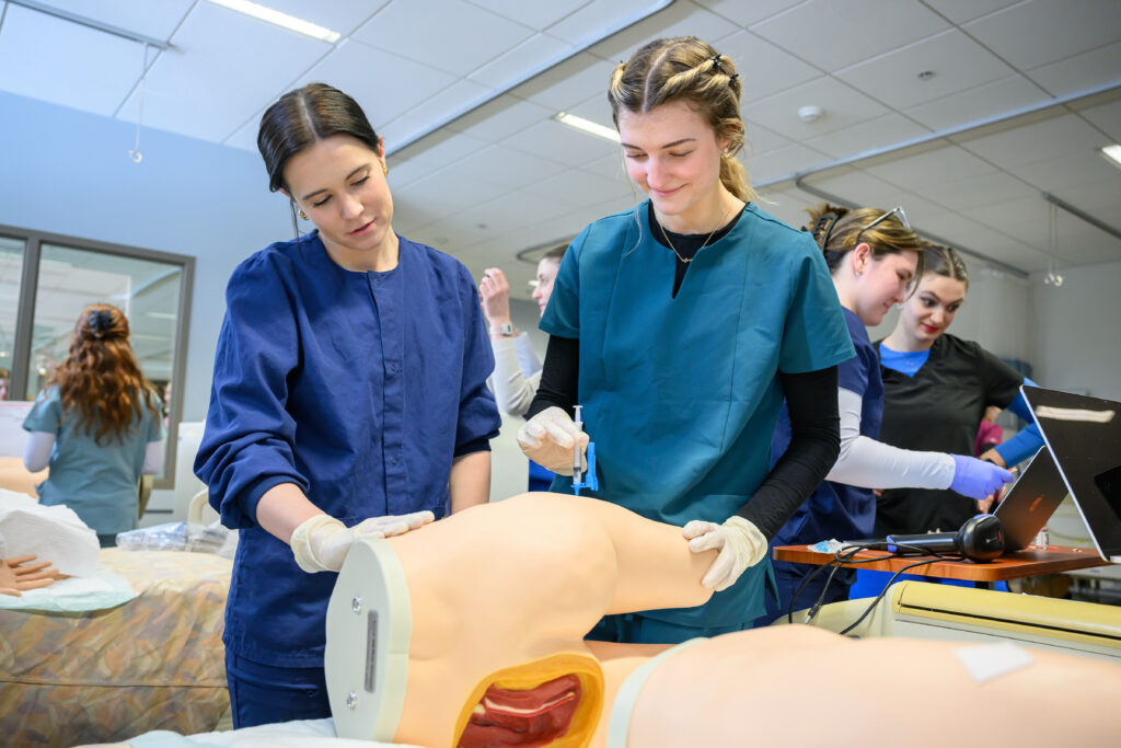 A WSU nursing student practicing an injection on a model while others work in the background.