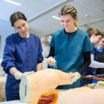 A WSU nursing student practicing an injection on a model while others work in the background.