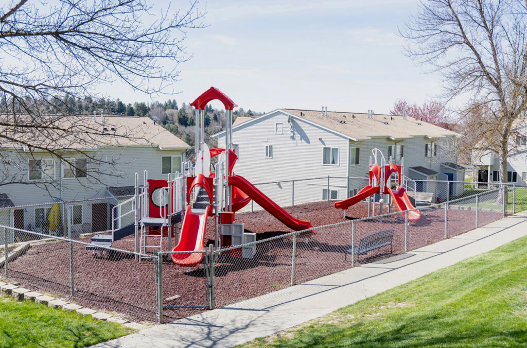 A playground area and play equipment, following a renovation, enclosed by a chain link fence near several apartments.