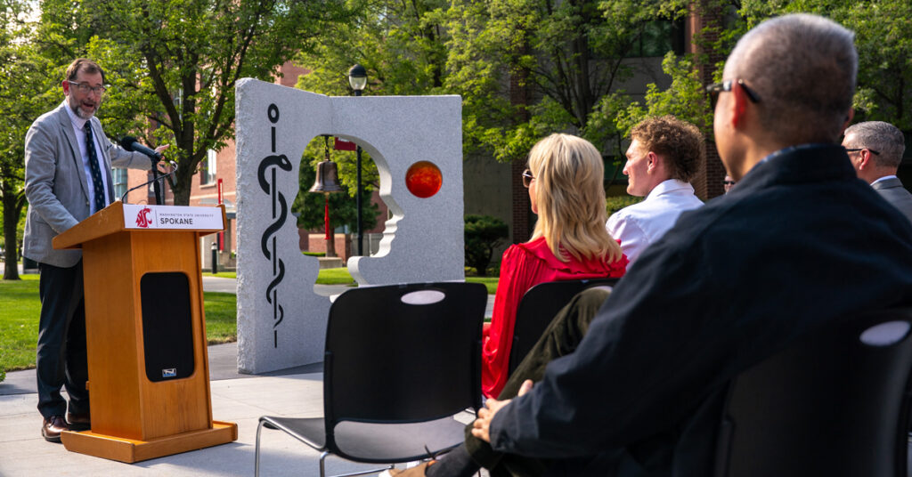 Chris Riley-Tillman gestures toward the Pages of Care sculpture from a podium during a dedication ceremony.