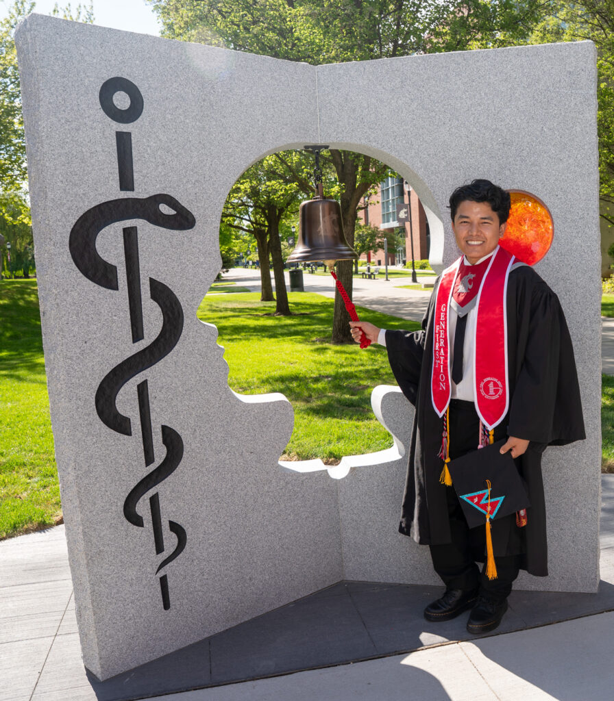 A WSU graduate in cap and gown ringing the bell in the Pages of Care sculpture.
