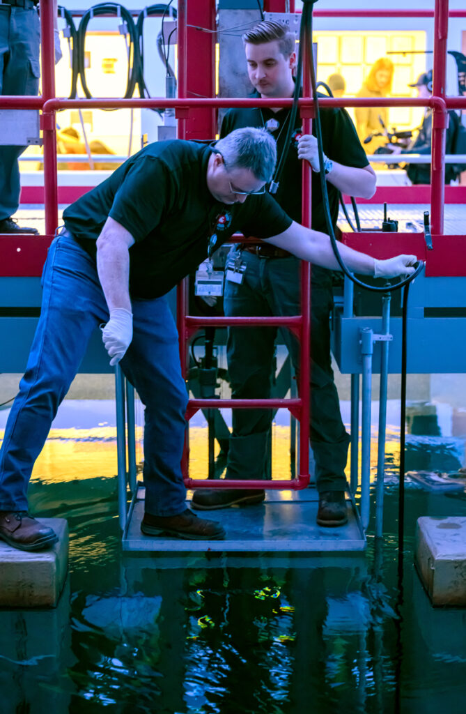 Two people on a platform lowering a device into the water inside a research nuclear reactor.