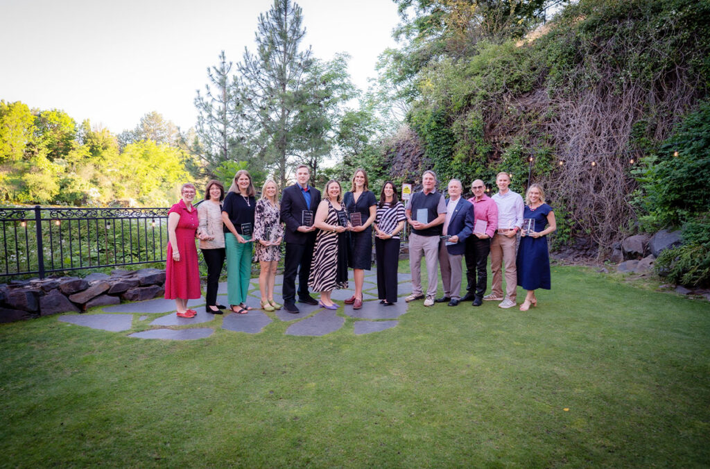 A group of award recipients stands on a landscaped lawn holding plaques with trees in the background.
