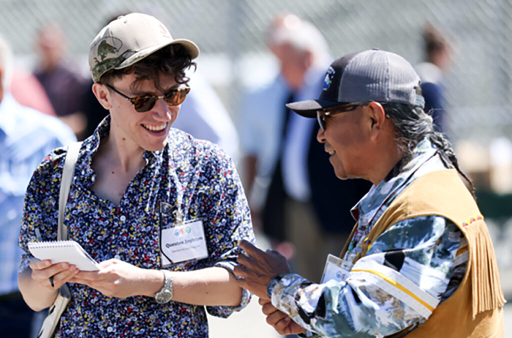 A reporter holds a pen and pad of paper while interviewing a tribal leader.