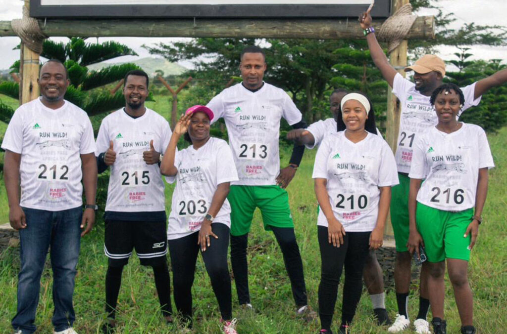 Seven participants wearing white "Run Wild! Rabies Free Africa 5K" event T-shirts and race bibs pose together outdoors on a grassy field beneath a large wooden sign structure. Several smile at the camera, with some giving thumbs-up or raising their arms in celebration.