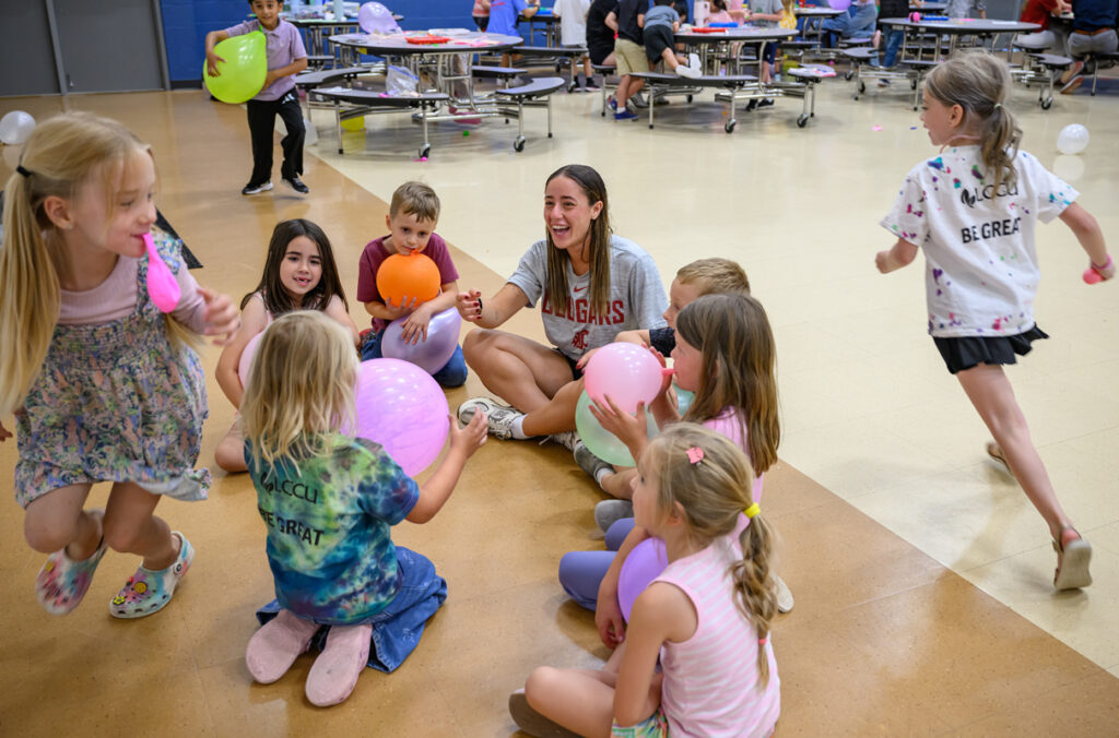 A WSU student-athlete sits in a circle with elementary school children holding balloons while two run children run around them.