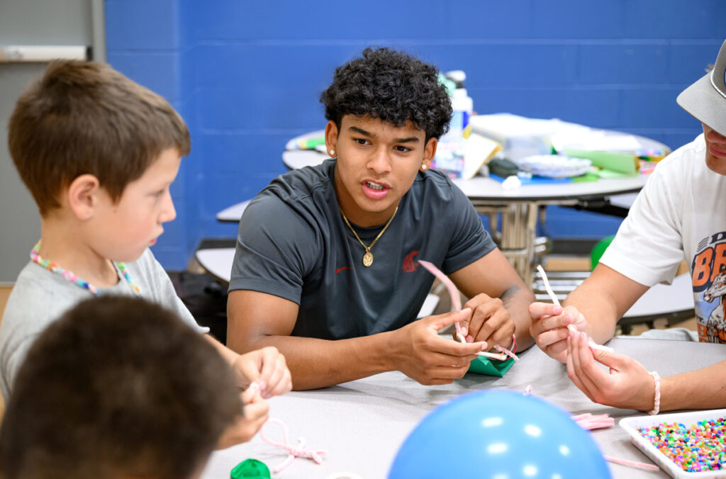 A WSU student-athlete sits at a craft table visting with elementary school children.