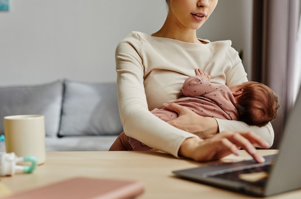 A mother breastfeeds an infant while working on a laptop