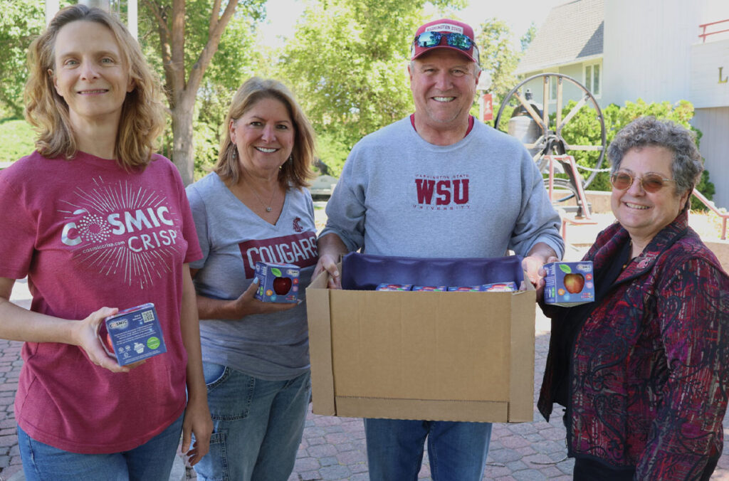 Four people wearing Coug attire stand in a line. The man in the middle holds a box of cosmic crisp apples.