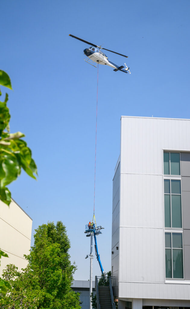Two workers on a boom lift guide a helicopter as it delivers a new LED light fixture to be installed.