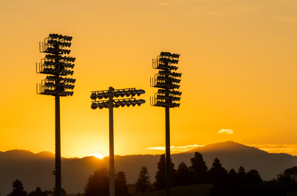The dark silhouette of three poles holding stadium lights against a yellow sky at sunrise.