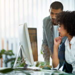 Two colleagues collaborate at a desk in a bright modern office — one sitting and one standing.