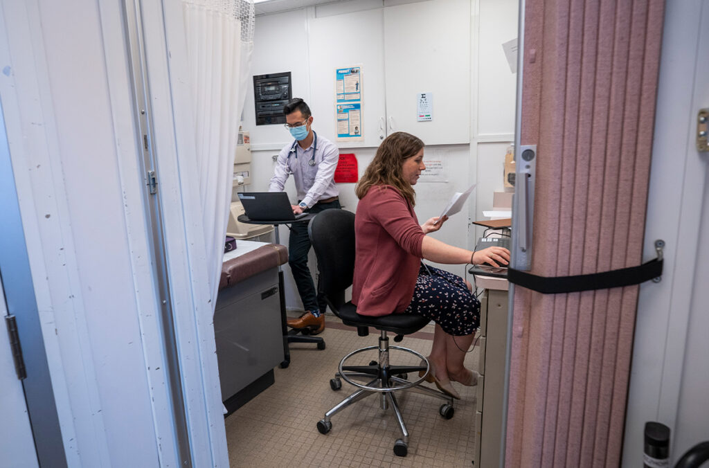 Healthcare staff work at computers inside a compact mobile clinic exam room, with medical equipment and an exam table visible in the background.