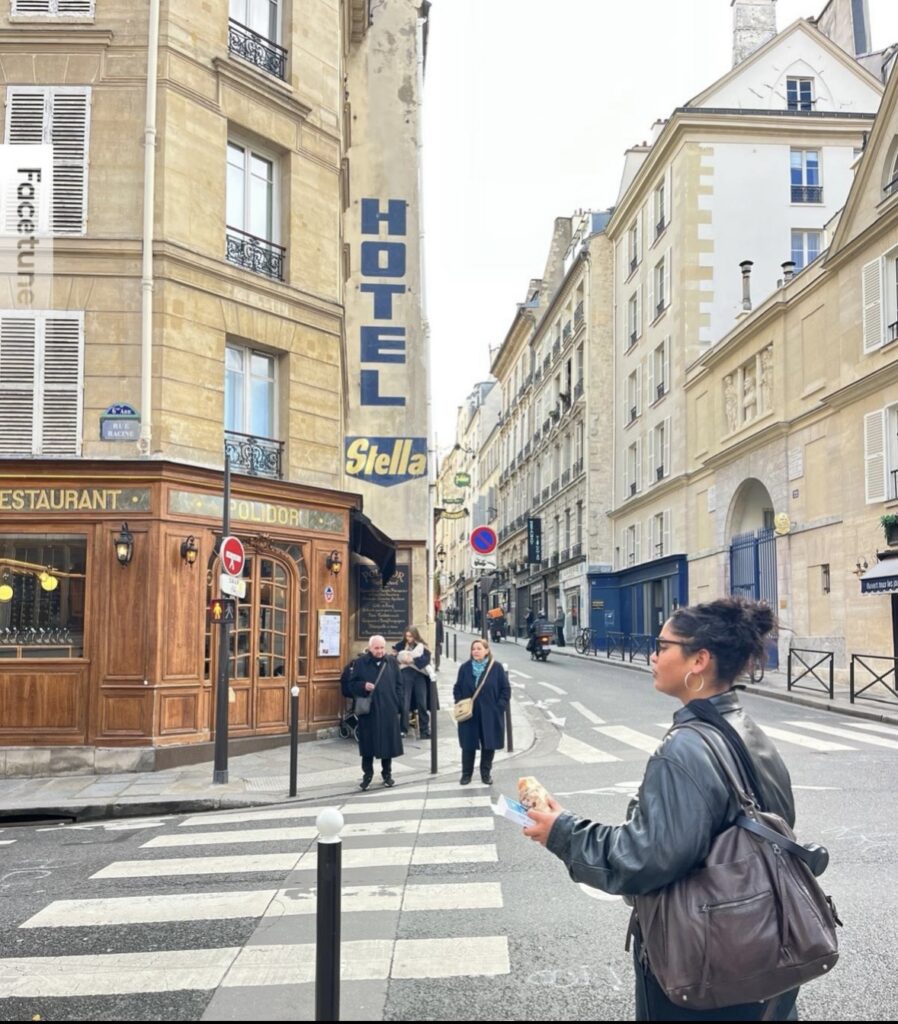 A street scene in Paris shows a corner restaurant with wood-paneled windows and a vertical blue HOTEL sign. The restaurant facade includes the text RESTAURANT, Stella, and POLIDOR. A blue street plaque reads RUE RACINE. Several pedestrians stand near a zebra crossing, while traffic signs, bollards, parked bicycles, and classic cream-colored buildings line the street.