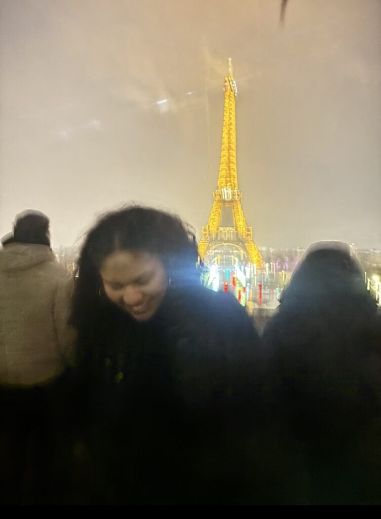 A nighttime view of the illuminated Eiffel Tower appears beyond a silhouetted crowd. The tower is framed between people in dark coats, with blurred lights and reflections across the scene.