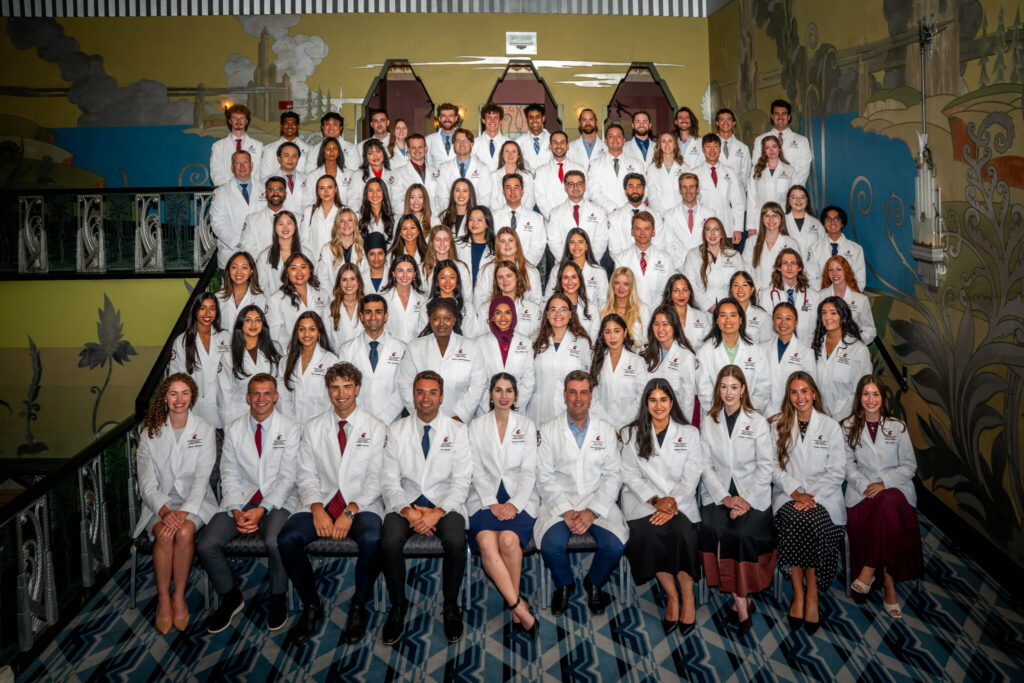 A large group of medical students in white coats poses for a class portrait on the stairs outside an auditorium following a White Coat Ceremony.
